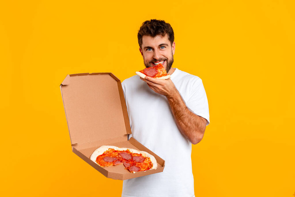 Az éhes európai fiatalember beleharap egy szelet pizzába, hangsúlyozva a kényelemevés és a gyorskajafogyasztás fogalmát, sárga alapon állva, kamerába nézve. Táplálkozási szokások - Fotó, kép