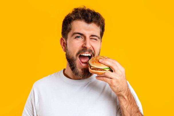 Mangeur drôle gars mordre un hamburger et clin d'oeil à la caméra sur fond jaune en studio. Portrait de jeune homme affamé mange délicieux cheeseburger. Nutrition, concept de repas Cheat - Photo, image