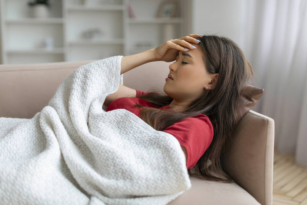Mujer asiática agotada con la mano en la frente, acostada en el sofá debajo de la manta, mujer coreana enferma que muestra signos de enfermedad o fiebre, relajándose en el cómodo entorno de la sala de estar, primer plano - Foto, imagen