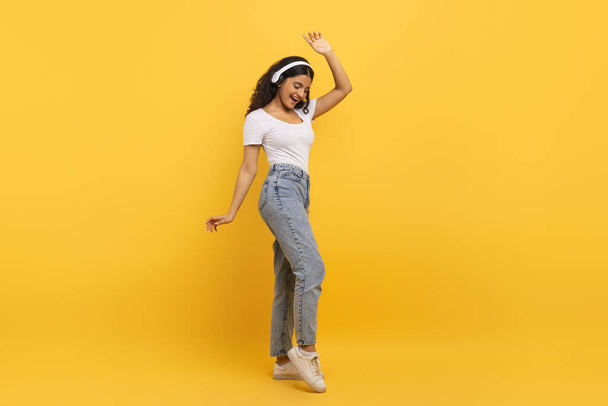 Щаслива молода студентка-індійка, яка носить повсякденний одяг за допомогою бездротових навушників і танцює із закритими очима, ізольована на жовтому студійному фоні, копіює простір, повна довжина - Фото, зображення
