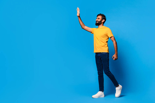Bearded indian man in yellow shirt walking and cheerfully waving hello, looking to the side at free space, full of energy on striking blue background - Photo, Image