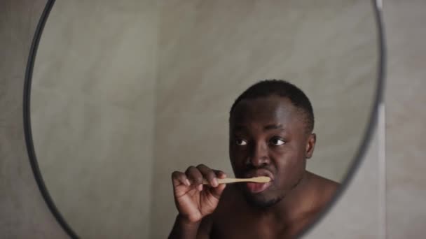 Keskipitkä lähikuva peili heijastus nuori lihaksikas Afrikkalainen amerikkalainen mies leikattu hiukset ja paljain rinnassa, seisoo kylpyhuoneessa aamulla ja harjaamalla hampaat valkoisella vaahtoava hammastahna - Materiaali, video