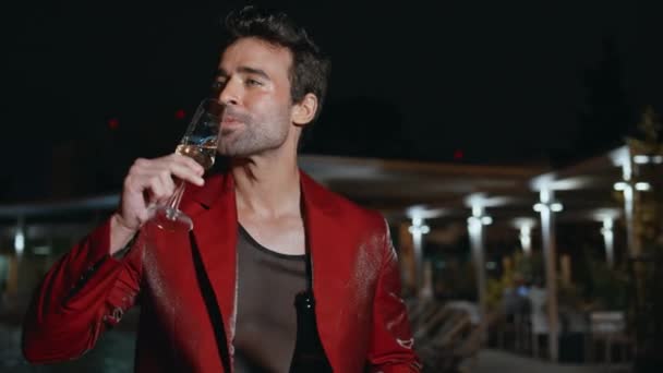 Party boy drinking sparkling wine at late evening hangout at poolside closeup. Excited hispanic man holding bottle exclusive alcohol relaxing at weekend night. Happy guy in glamorous red suit chilling - Footage, Video