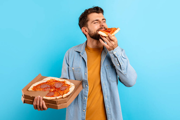 Голодный человек тысячелетия наслаждается вкусом пиццы ломтик холдинг открытую коробку доставки на синий студийный фон, концепция переедания и удовольствие от обмануть еды во время диеты - Фото, изображение