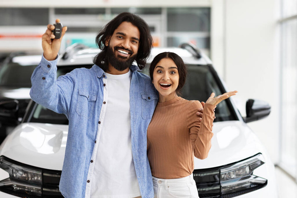 Opgewonden duizendjarig indiaans paar naast nieuwe witte auto, met de sleutel van gloednieuwe luxe auto 's en gebaren, vieren aankoop bij autodealer - Foto, afbeelding