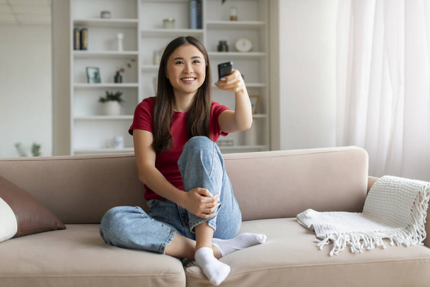 Boldog fiatal ázsiai nő távirányítóval a kezében tv-t néz otthon, vidám koreai nő élvezi televíziós show, pihentető kanapén kényelmes nappali belső, másolás tér - Fotó, kép
