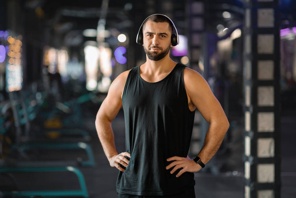 Πορτρέτο του όμορφος νεαρός μυώδης άνδρας στα ακουστικά Ποζάροντας στο γυμναστήριο, χαμογελώντας αυτοπεποίθηση Fitness Trainer στέκεται στο σύγχρονο αθλητικό σύλλογο Εσωτερικών και κοιτάζοντας κάμερα, Διαφήμιση Υγιεινός τρόπος ζωής - Φωτογραφία, εικόνα