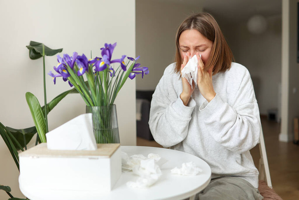 Sick young woman suffering from allergy, fever or flu blows nose and sneezing in paper tissues, sits by table at home kitchen. Girl with runny nose, pollen allergy, flu, cold, rhinitis, sinusitis - Photo, Image