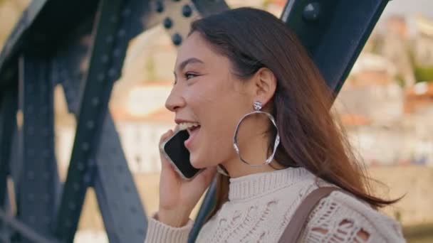 Χαρούμενη μελαχρινή μιλάει κελί μοιράζονται καλά νέα στο κλείσιμο σιδερένια γέφυρα. Ευτυχισμένο μεξικάνικη κυρία καλώντας smartphone χαρά στο περπάτημα το Σαββατοκύριακο. Χαμογελώντας μαύρο μοντέλο μαλλιών μιλώντας τηλέφωνο. Ενθουσιασμένη γυναίκα σε εφημερία - Πλάνα, βίντεο