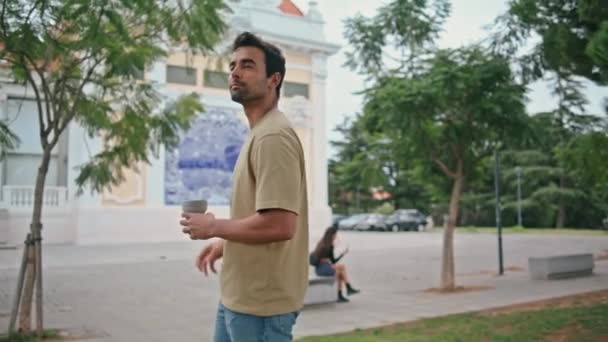Latin guy looking town area at weekend walk. Handsome unshaven man strolling city street holding coffee cup takeaway at summer vacation. Relaxed happy male tourist exploring urban district alone. - Footage, Video