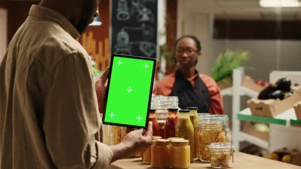Client afro-américain tenant tablette avec mise en page écran vert dans un magasin bio local, regardant chromakey à côté de produits sans additifs. Le client régulier utilise un appareil avec un espace de copie isolé. Caméra A. - Séquence, vidéo