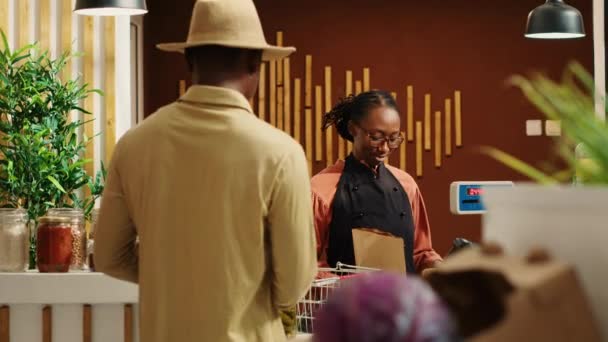 Africký americký prodejce pracující u pokladny v supermarketu, váží organické přídatné látky bez ovoce a zeleniny. Prodejce prodává čerstvé bio produkty mužskému zákazníkovi na pokladně. Fotoaparát A. - Záběry, video