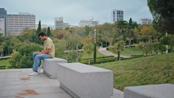Rento kaveri tekstailee kännykkäviestiä betonipenkillä. Rento italialainen mies messaging älypuhelimella istuu vihreä kaupunginpuisto yksin. Huoleton tuhatvuotinen hipster käyttää matkapuhelinta viestintään. - Materiaali, video