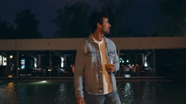 Latin man chilling at night walk near water pool. Carefree handsome guy holding glass with alcohol relaxing at city outdoors party. Relaxed unshaven hipster moving at sound enjoy evening weekend rest. - Footage, Video