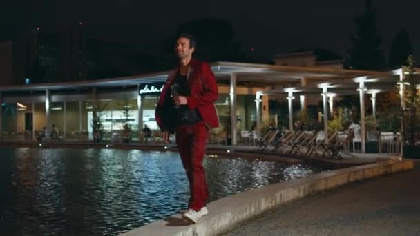 Trendy hipster genieten van 's nachts feest wandelen aan het zwembad. Gelukkig zorgeloze man in glamoureuze rode pak met champagne glas ontspannen in het zwembad club. Glimlachende Spaanse man die laat in de avond aan het chillen is. - Video