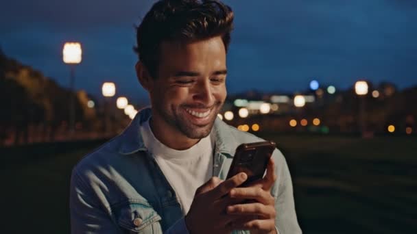 Laughing man answering message at smartphone resting in night city close up. Happy italian guy texting cell phone in dark town street. Unshaven handsome male chatting in social networks outdoors. - Footage, Video