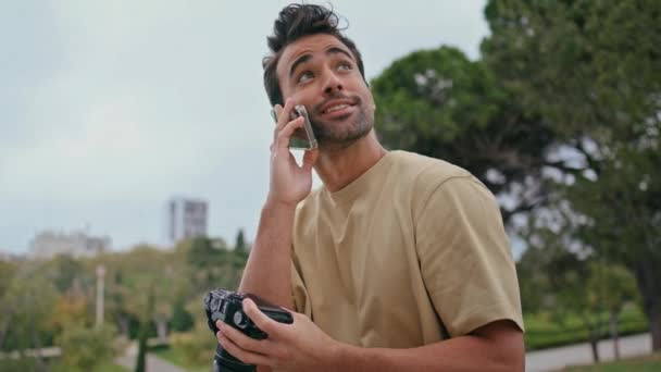 Boldog riporter beszél okostelefon keres fotózás professzionális kamera zöld parkban közeli. Jóképű spanyol férfi telefonál és fotókat néz. Mosolygó fotós élvezze a beszélgetést. - Felvétel, videó