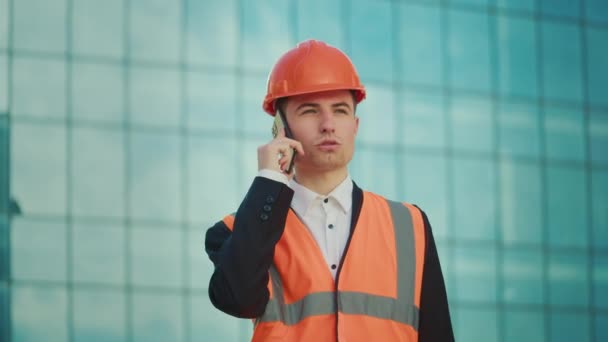 Engenheiro masculino ou arquiteto vestindo um capacete laranja e jaqueta de segurança conversando no telefone, segurando documentos de pé perto de um edifício com janelas azuis - Filmagem, Vídeo