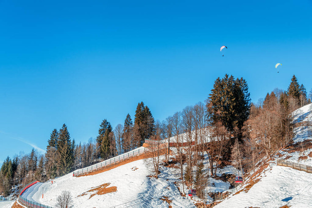 Uma paisagem serena de inverno, exibindo uma colina coberta de neve com árvores nuas e dois parapentes no céu azul claro. Ideal para conceitos de aventura, natureza e tranquilidade - Foto, Imagem