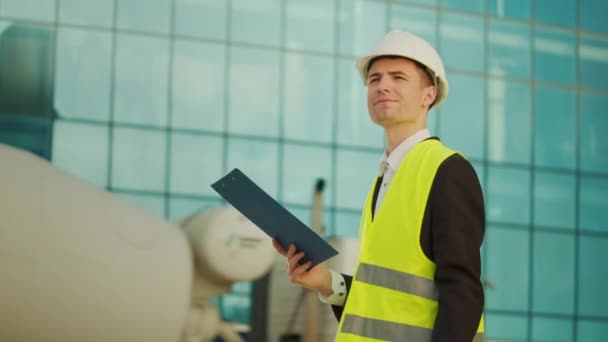 Mężczyzna inżynier lub architekt noszący w hełmie i kurtce bezpieczeństwa, patrząc rysunki budowlane w jego ręce chodzenia w pobliżu budynku z niebieskimi oknami - Materiał filmowy, wideo