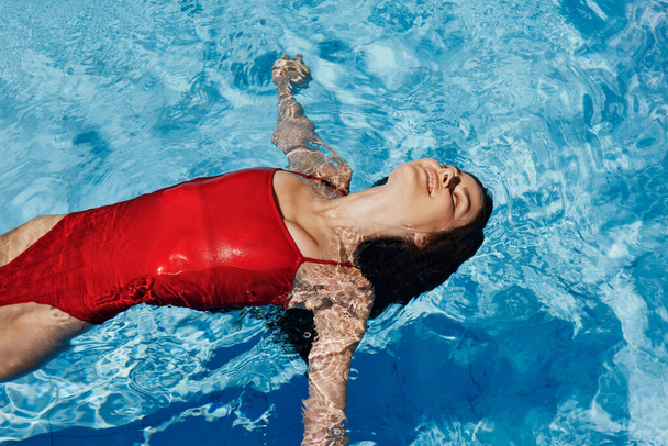 Счастливая женщина плавает в бассейне в красном купальнике с рыжими длинными волосами, защита кожи солнцезащитным кремом, концепция отдыха в отпуске. Высокое качество фото - Фото, изображение