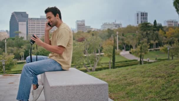 Latin fotós beszél okostelefon zöld parkban. Boldog gondtalan fickó hívja okostelefon keres képeket professzionális fényképezőgép ülő pad. Mosolygós ember turista megosztani utazási benyomás telefonon. - Felvétel, videó