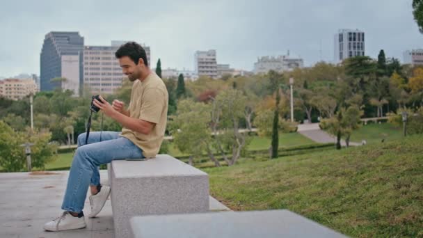 Profesjonalny fotograf odbierający telefon na ławce w parku betonowym. Szczęśliwy Włoch mówi smartfon patrząc na ekranie aparatu fotograficznego na zewnątrz. Uśmiechnięty turysta mówiący przez komórkę oglądający fotografię. - Materiał filmowy, wideo