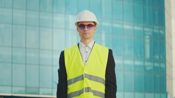 Portrét úspěšného mladého muže-inženýra nebo architekta v přilbě a bezpečnostní vestě, držící dokumenty stojící poblíž budovy s modrými okny - Záběry, video