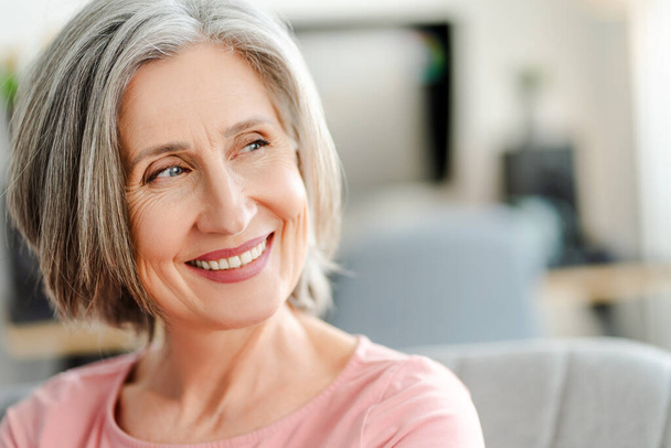Closeup portret glimlachend zelfverzekerde oudere vrouw zitten op comfortabele bank thuis. Happy gepensioneerde vrouw met stijlvol grijs haar, witte tanden wegkijkend. Natuurlijke schoonheid, gezonde levensstijl concept  - Foto, afbeelding