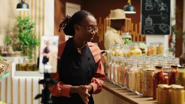 Vendedor afroamericano promoviendo salsas caseras y miel, la creación de anuncios de vídeo en línea para presentar productos ecológicos en el supermercado de residuos cero. Mujer grabando anuncio promocional. Cámara A. - Imágenes, Vídeo