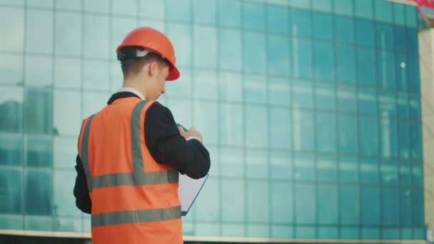 Ingeniero o arquitecto masculino con un casco naranja y una chaqueta de seguridad hablando por teléfono, sosteniendo documentos de pie cerca de un edificio con ventanas azules - Metraje, vídeo