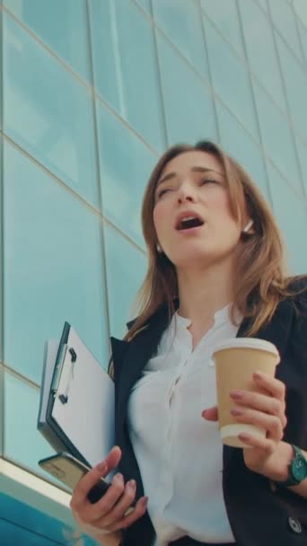 Die Geschäftsfrau trägt einen formellen Anzug mit Kopfhörern und telefoniert mit einer Kaffeetasse, während sie in der Nähe eines modernen gläsernen Geschäftsviertels spaziert. Kaffeepause im Freien - Filmmaterial, Video