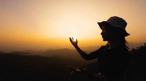 Силуэт Молодая женщина молится на горе, руки протянуты наблюдая красивый драматический восход солнца.   - Фото, изображение