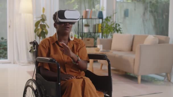 Medium long shot di donna afroamericana a sedia a rotelle che utilizza occhiali VR durante la riproduzione di videogiochi a casa - Filmati, video