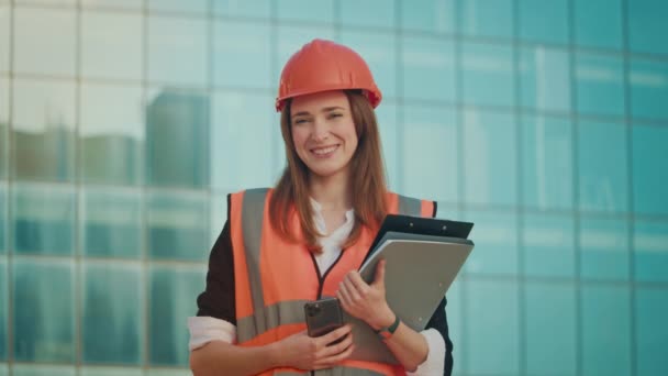 Başarılı Genç Bir Kadın Mühendis veya Mimar Portresi Turuncu Miğfer ve Güvenlik Ceketi Giyiyor, Elinde Mavi Pencereli Bir Binanın Yanında Duran Belgeler Tutuyor - Video, Çekim