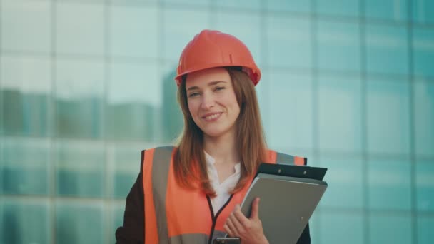 成功した若い女性エンジニアまたは建築家の肖像画 オレンジヘルメットと安全ジャケットを着用し,ブルーウィンドウで建物の近くに立っている文書を保持 - 映像、動画