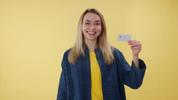Portrét usměvavé ženy držící a dívající se na plastovou kreditní kartu uvnitř. Emocionální běloška, která provádí online platbu moderními technologiemi. Izolované přes žluté pozadí studia. - Záběry, video
