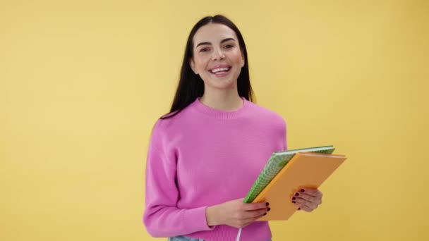 Positive female freshman in cool outfit showing books and smiling while standing over yellow studio background. Intelligent brunette woman enjoying studying and achieving new skills and knowledge. - Footage, Video