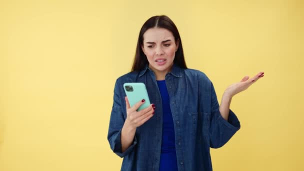 Valkoihoinen nainen kohauttaa olkapäitä ja tunne tyytymätön katsellessaan mobiili näyttö keltaisella taustalla. Surullinen nainen brunette ottaa ongelmia internet-yhteyden moderni älypuhelin. - Materiaali, video