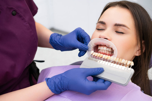 De tandarts selecteert zorgvuldig de glazuur schaduw voor de jonge brunette vrouwen tanden voor het begin van de whitening procedure. - Foto, afbeelding