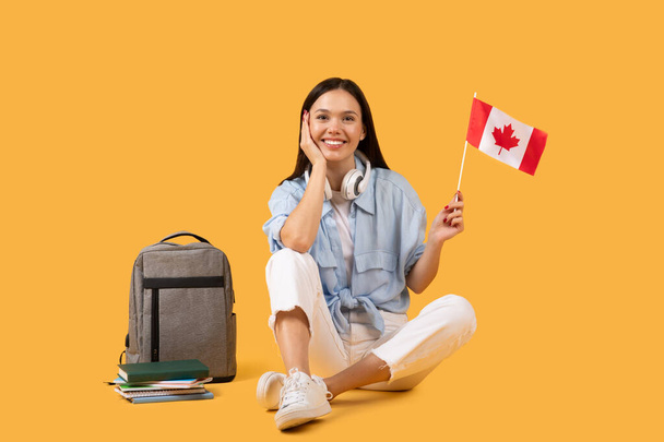 Felice studentessa che tiene la bandiera canadese, indossa le cuffie, seduta con posa rilassata accanto a libri e zaino sullo sfondo giallo - Foto, immagini