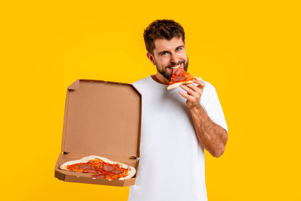 Ευτυχισμένος καυκάσιος άνθρωπος απολαμβάνοντας φέτα πίτσα από το κουτί παράδοσης, ενδίδει σε εξαπατήσει γεύμα, απολαμβάνοντας πρόχειρο φαγητό στο στούντιο ρύθμιση πάνω από κίτρινο φόντο, χαμογελώντας στην κάμερα. Γρήγορη κατανάλωση τροφίμων - Φωτογραφία, εικόνα