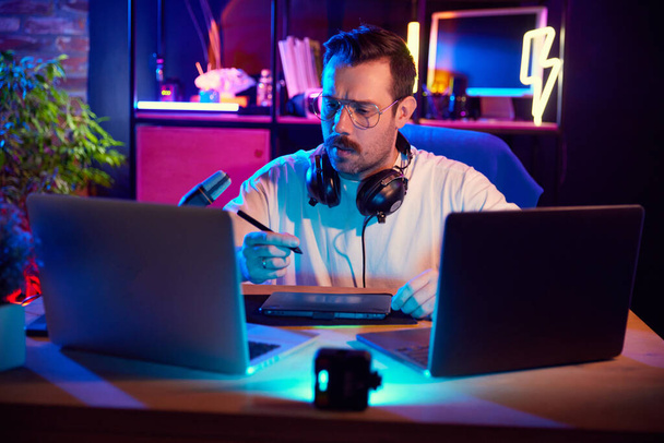 Νεαρός άνδρας με ακουστικά σε neon lit σπίτι-στούντιο που εργάζονται στον υπολογιστή δημιουργώντας περιεχόμενο για το blog του. Έννοια των νέων με τα μέσα κοινωνικής δικτύωσης και έξυπνη εργασία, τρόπος ζωής, σε απευθείας σύνδεση πλατφόρμα streaming. - Φωτογραφία, εικόνα