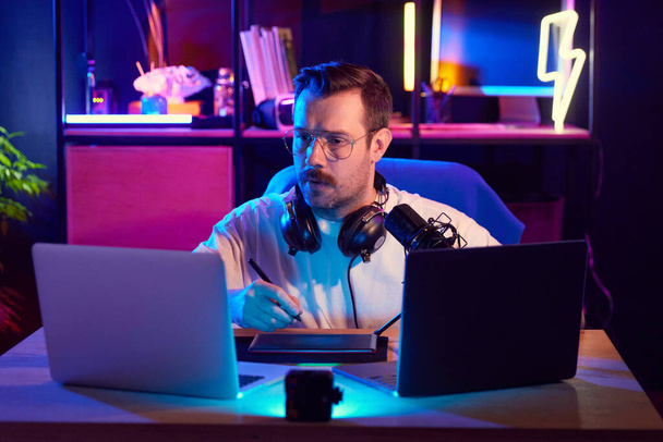 Hombre enfocado en auriculares y gafas podcasting y mirando a la computadora portátil, rodeado de luces de neón en el hogar-estudio. Concepto de jóvenes con redes sociales y trabajo inteligente, estilo de vida, streaming. - Foto, imagen