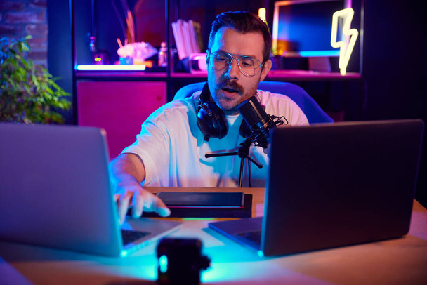 Man in vibrant studio setting speaking into microphone and typing on laptop keyboard in neon light at home-studio. Concept of youth people with social media and smart working, lifestyle, digital. - Photo, Image