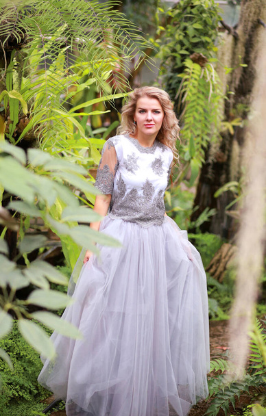 Una hermosa rubia en un elegante vestido de tul en un jardín botánico. Fondo verde y plantas tropicales. Concepto de belleza y moda - Foto, imagen