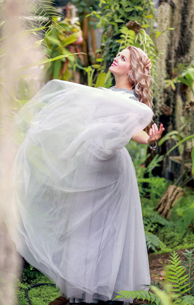 Una hermosa rubia se gira en un elegante vestido de tul en un jardín botánico. Fondo verde y plantas tropicales. Concepto de belleza y moda - Foto, imagen