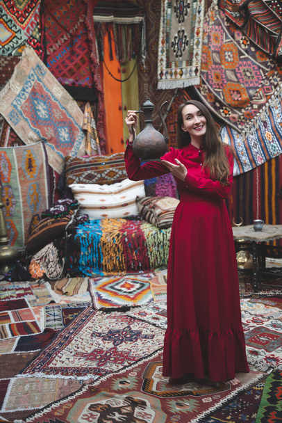 hermosa chica en un vestido rojo con una tetera en un interior tradicional turco con muchas alfombras en la pared - Foto, imagen