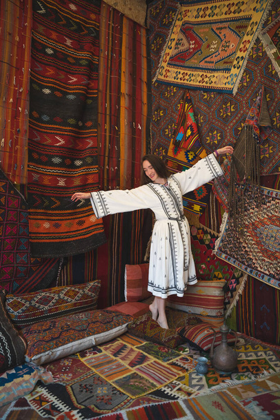 piękna dziewczyna w białej sukience z czajniczek w tradycyjnym tureckim wnętrzu z wieloma dywanami na ścianie - Zdjęcie, obraz