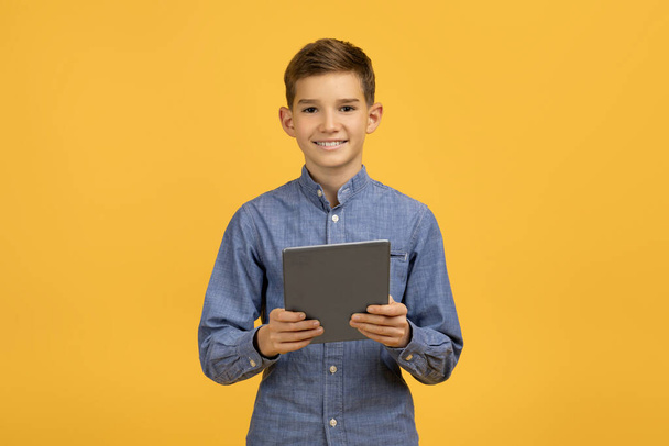 Joyeux adolescent tenant une tablette numérique dans les mains et souriant à la caméra, joyeux garçon debout sur fond jaune vif, indiquant l'utilisation de la technologie et des possibilités d'apprentissage en ligne - Photo, image